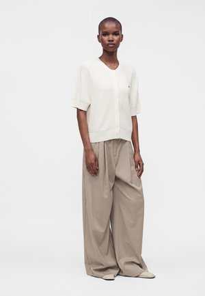Personne portant un haut blanc à manches courtes boutonné et un pantalon ample beige à jambes larges, debout devant un fond blanc uni.