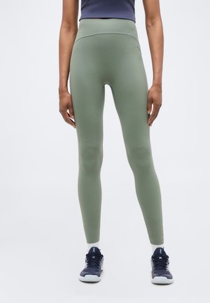 Isik seisab, kandes kõrge vöökohaga helerohelisi retuuse, tumesiniseid tossusid valgete talladega ja tumedat lühikest särk, tavalise tausta ees.