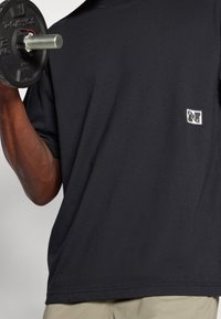 Levý loket drží 5 kg hmotnostní kotouč blízko ramene, oblečený v černém tričku s krátkým rukávem značky Nike a béžových kalhotách.