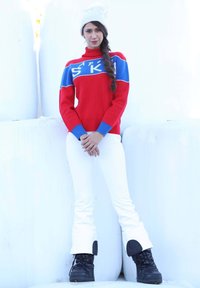 Maglione rosso lavorato a maglia con dettagli blu e testo "SKI", abbinato a pantaloni bianchi e stivali invernali neri. Capelli intrecciati e un berretto bianco.
