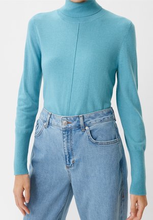 Ljusblå stickad tröja med hög krage i kombination med high-waisted ljusa jeans på en person mot en enhetlig bakgrund.