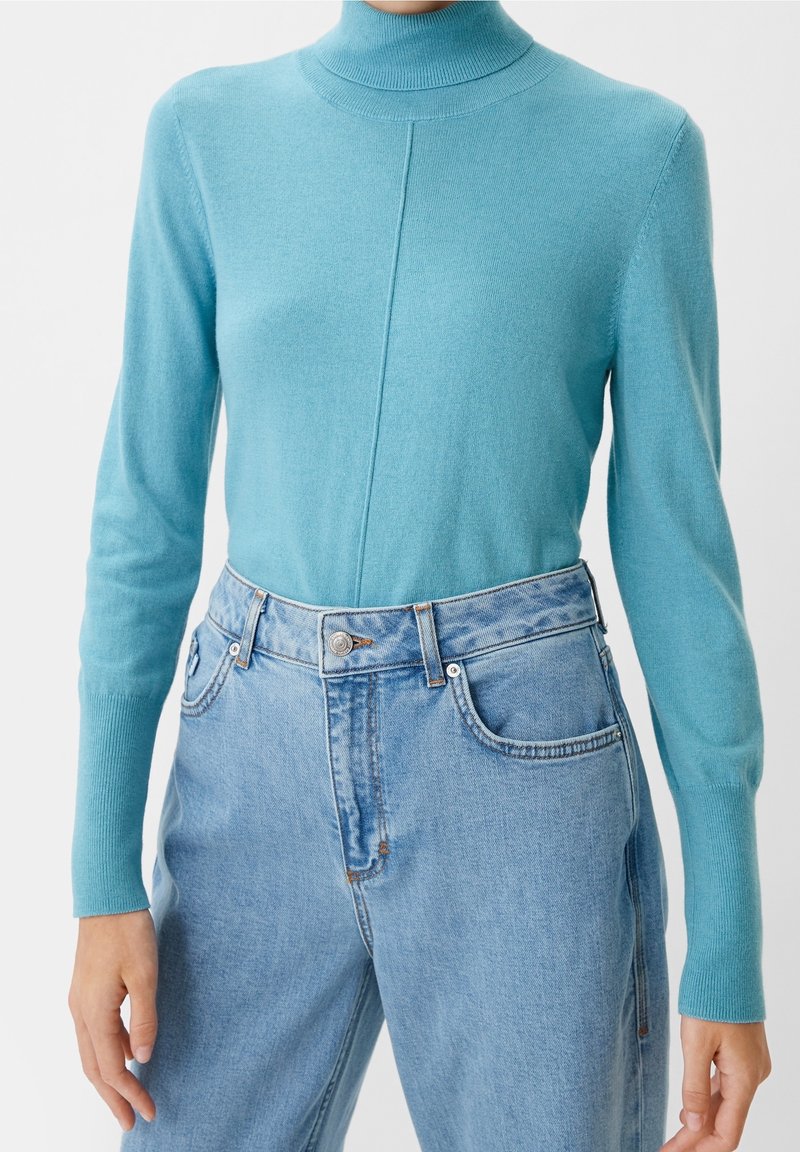 Suéter de cuello alto azul claro combinado con unos vaqueros de cintura alta de lavado claro en una persona frente a un fondo liso.