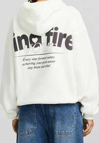 Sudadera blanca con capucha, con gran texto negro en la parte posterior, que presenta frases motivacionales y un símbolo de corazón. Tejido suave, corte relajado.