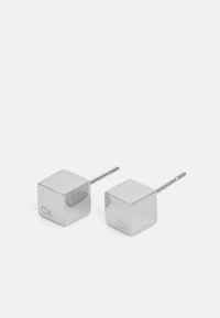 Hopeakubehelmikorvakorut kiiltävällä pinnalla, jossa on neppikloosi ja toisella puolella kaiverrettu "CK". Minimalistinen muotoilu, geometrinen muoto.