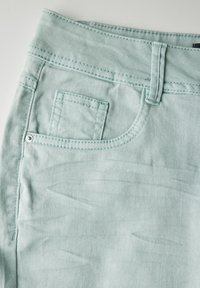 Tela de mezclilla azul claro con una textura suave, que presenta un pequeño bolsillo frontal, pasadores para cinturón y detalles de costura visibles.