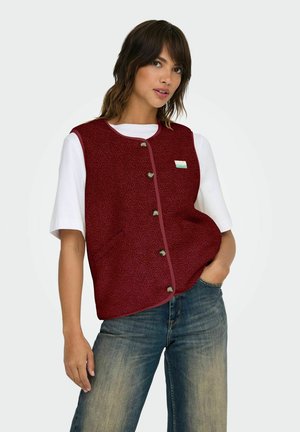 Een vrouw in een knoop-jack met textuur in bordeauxrood over een wit shirt met korte mouwen, met haar linkerhand in de zak van haar blauwe spijkerbroek, tegen een effen achtergrond.