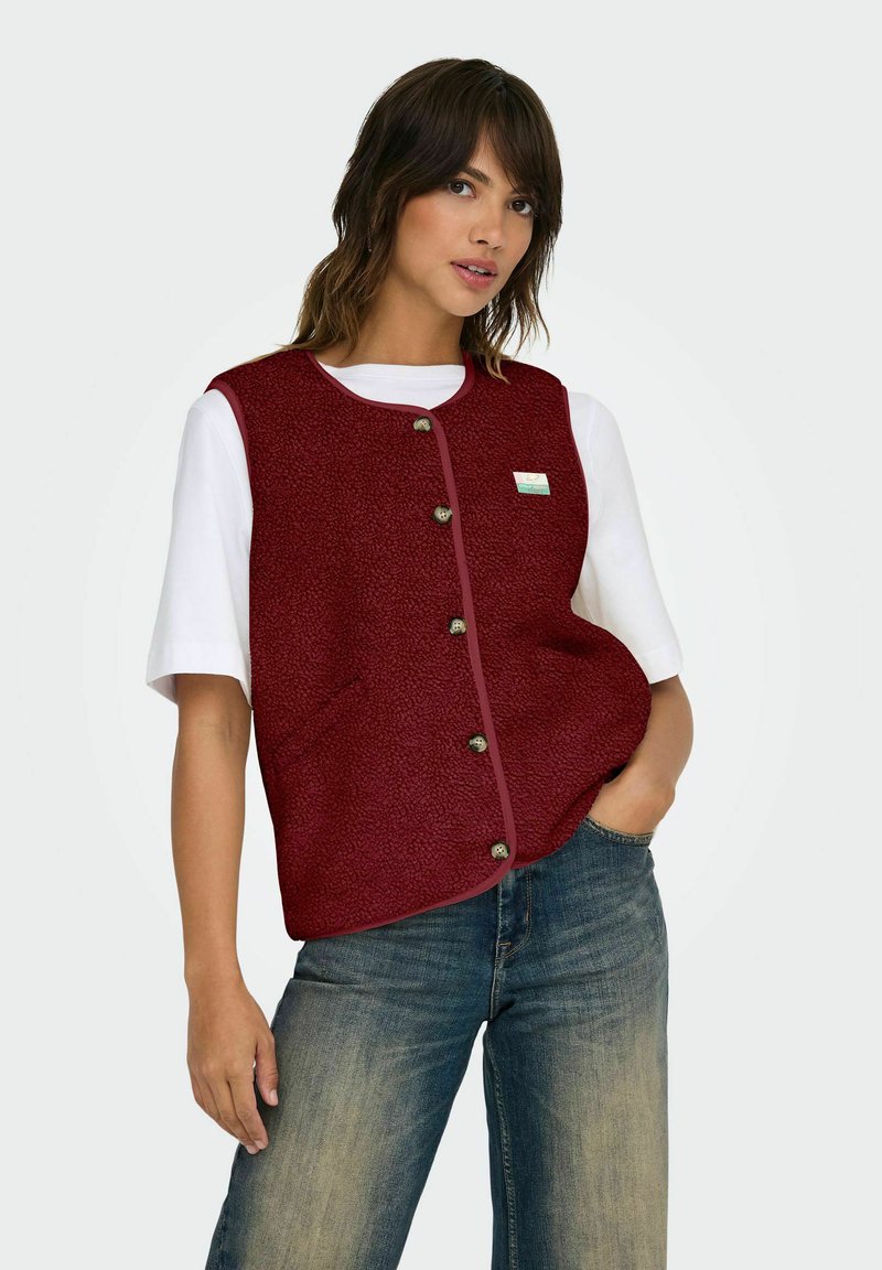 Een vrouw in een knoop-jack met textuur in bordeauxrood over een wit shirt met korte mouwen, met haar linkerhand in de zak van haar blauwe spijkerbroek, tegen een effen achtergrond.