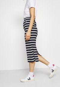 Person i vit t-shirt, marinblå och vit randig midi kjol, vita strumpor och vita sneakers med svarta och röda detaljer, som går.