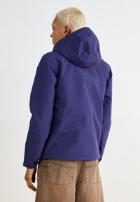 Veste à capuche violette avec un extérieur lisse, cordon de serrage ajustable à l'ourlet et coupe décontractée. Associée à un pantalon large marron clair.