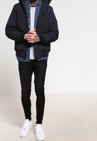 Marine bomberjakke med hætte, lagd oven på en lysegrå t-shirt og en blåternet skjorte. Sorte slim-fit jeans og hvide sneakers fuldender looket.