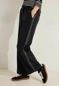 Zwarte wijde broek met zijzakken, voorzien van een leren streep langs het been, gecombineerd met zwarte sneakers en witte veters.
