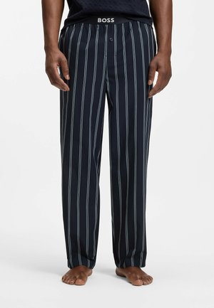 Homme portant un pantalon bleu marine avec des rayures blanches verticales et une ceinture noire portant l'inscription "BOSS", debout pieds nus sur fond blanc.