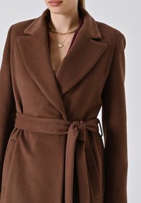 Bruine wikkeljas van textiel met een structuurbewerking, met een brede kraag en een strikceintuur in de taille. Twee zichtbare zakken aan de voorkant.