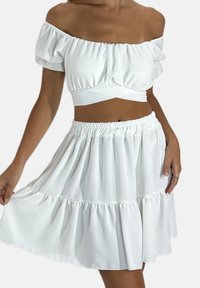 Tenue blanche composée de deux pièces : un crop top épaules dénudées avec des détails froncés et une jupe évasée à ourlet volanté. Texture de tissu douce.