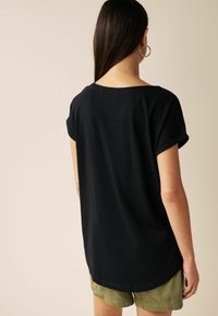 Zwarte shirt met korte mouwen, omgeslagen mouwen, losse pasvorm, ronde zoom, gemaakt van zachte stof, gedragen over lichtgroene shorts.