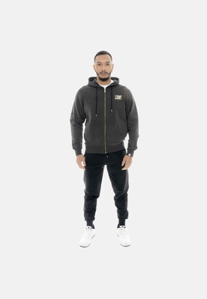 Homme debout portant un sweat à capuche noir zippé avec des détails dorés, un pantalon noir et des baskets blanches sur un fond clair uni.