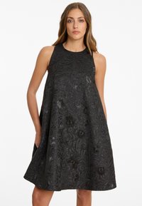 Femme portant une robe noire sans manches avec un motif floral texturé, se tenant avec une main dans la poche devant un fond blanc.