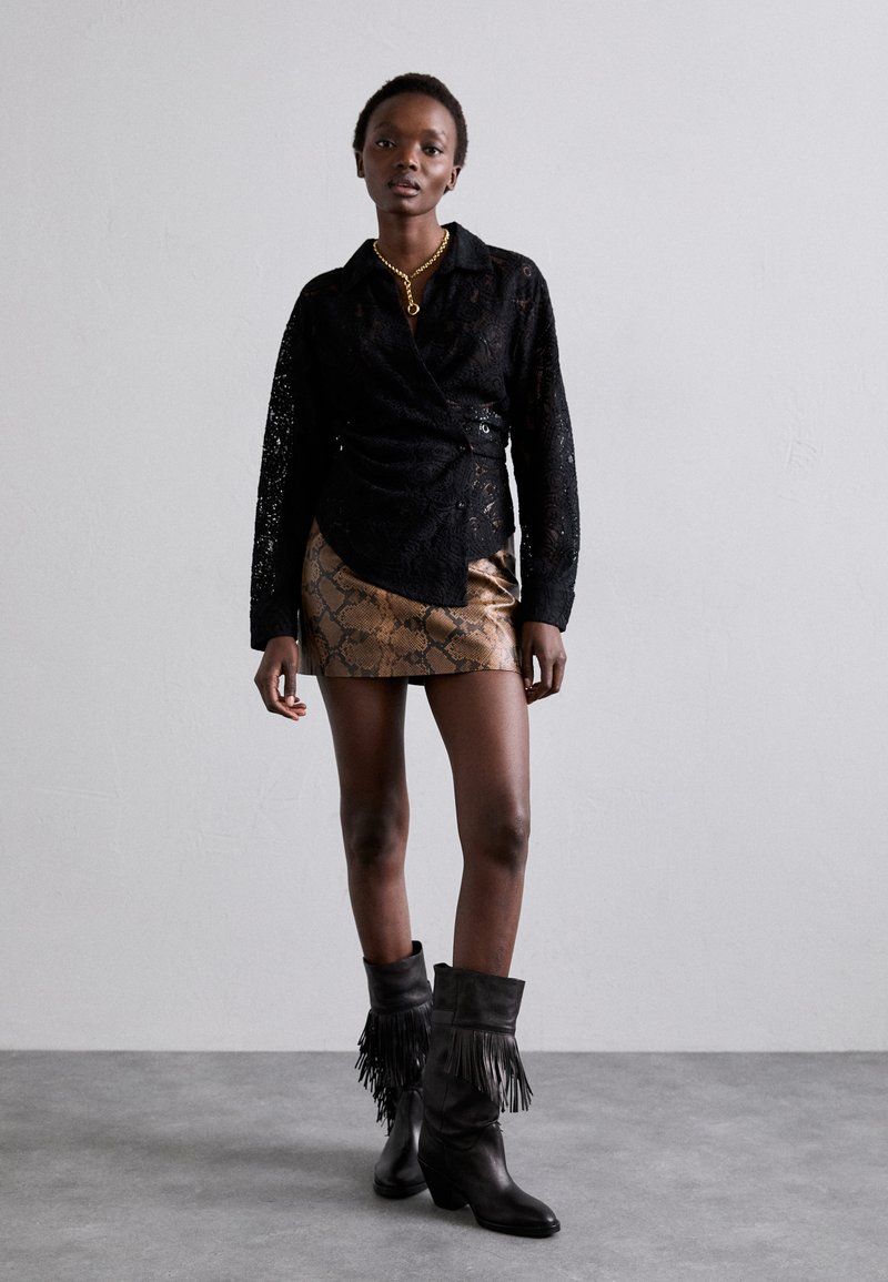 Woman wearing a black lace blouse, brown snakeskin mini skirt, black fringe boots, and a gold chain necklace standing against a plain gray wall.