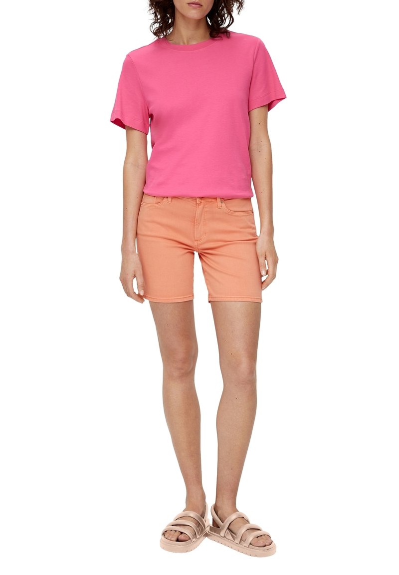 T-shirt rose à manches courtes en coton associée à un short en denim orange. Le modèle porte des sandales beiges à lanières, mettant en valeur une tenue décontractée d'été.