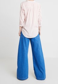 Persona che indossa una camicia a strisce verticali rosa e bianche con maniche a tre quarti e jeans blu a gamba larga, in piedi di spalle.
