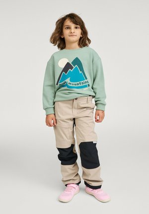 Dziecko o brązowych włosach do ramion ubrane w zielony sweter z grafiką gór, beżowe spodnie z czarnymi łatami oraz różowe trampki, stoi nieruchomo.