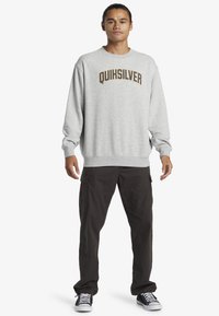 Sweatshirt gris clair avec le logo "QUIKSILVER" en marron foncé, associé à un pantalon cargo noir. Design décontracté, poignets et ourlet côtelés. Basket noires.