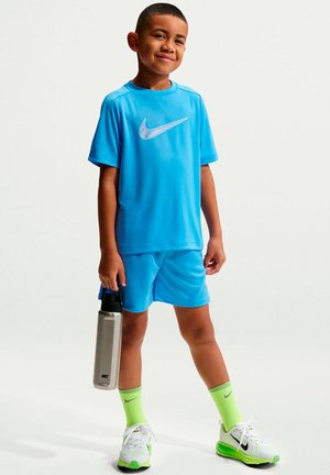 Jongen in blauwe Nike sportkleding en neon groene sokken, die een zilveren waterfles vasthoudt, staand en glimlachend.