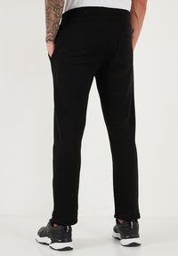 Schwarze Jogginghosen mit einem elastischen Bund, Seitentaschen und einer Gesäßtasche, aus weichem Stoff. Getragen mit schwarzen Sportschuhen.