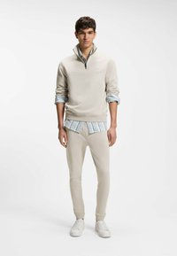 Νεαρός άνδρας φοράει μπεζ φούτερ με φερμουάρ μέχρι τη μέση πάνω από μπλε ριγέ πουκάμισο, μπεζ αθλητική φόρμα και άσπρα αθλητικά παπούτσια, στέκεται μπροστά σε λευκό φόντο.