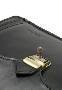 Kąt czarnej skórzanej torby z zapięciem w kolorze złotym i włożonym małym kluczykiem, widoczne szwy oraz wytłoczone nazwisko marki "WORS".