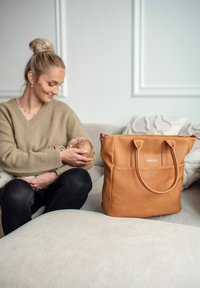 Kožená taška přes rameno hnědé barvy s texturovaným povrchem, vybavená dvěma pevnými uchy, přední kapsou a vyraženým logem. Položena na světlé látkové pohovce.