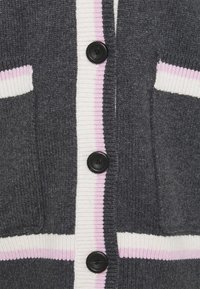Gros plan d'un cardigan en maille gris foncé avec des boutons noirs et une bordure rayée blanche et rose le long de la ligne des boutons, des poches et de l'ourlet.