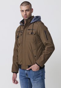 Braune gepolsterte Jacke mit grauem Kapuzen, Reißverschlusstaschen und Druckknopfverschluss. Verfügt über einen gerippten Bund und Saum.