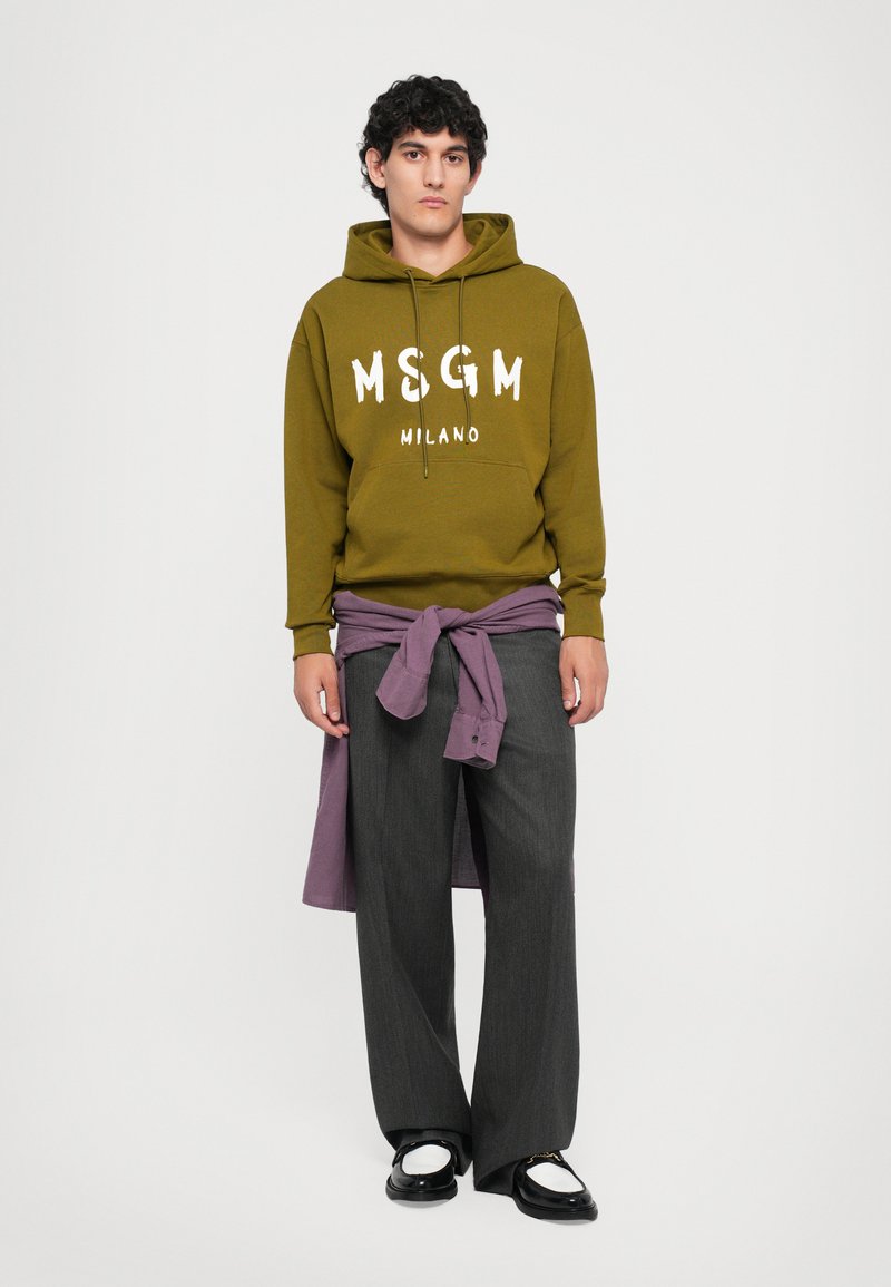 Jeune homme portant un sweat à capuche MSGM Milano vert olive, un pantalon large gris foncé, une chemise violette nouée autour de la taille et des chaussures noir et blanc.