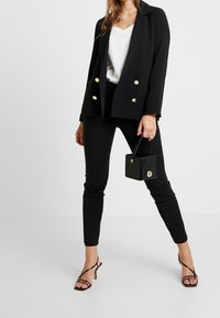 Černý dvouřadý blazer se zlatými knoflíky, v kombinaci s těsně padnoucími černými kalhotami a sandály na vysokém podpatku. Drží malou černou kabelku s krokodýlím vzorem.
