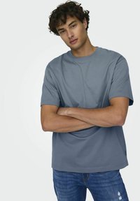 Lichtblauw katoenen T-shirt met korte mouwen en een ronde halslijn. Het model dat het draagt heeft zijn armen over elkaar tegen een eenvoudige achtergrond.