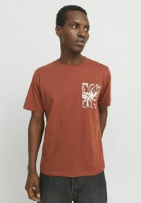 T-shirt kratkih rukava rust boje, izrađen od teksturirane tkanine, s džepom s lijeve strane s bijelim apstraktnim uzorkom.