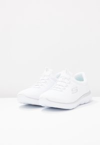 Vita atletiska skor med en stickad ovandel, snörning och en texturerad, dämpad sula. Silverdetaljer nära hälen och logotyp.