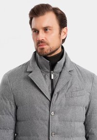 Abrigo acolchado gris con cuello, con cierre de cremallera y botones en la parte delantera. Está hecho de una tela texturizada con detalles sutiles y un bolsillo en el pecho.