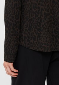 Camisola de manga longa em castanho-escuro com um padrão de estampado de leopardo preto. Tecido macio, barra arredondada e mangas com punhos. Acentuada com costura contrastante.