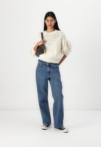 Pull en maille ivoire avec des détails ajourés, manches bouffantes, associé à un jean droit bleu et des baskets noires. Petit sac gris porté à l'épaule.