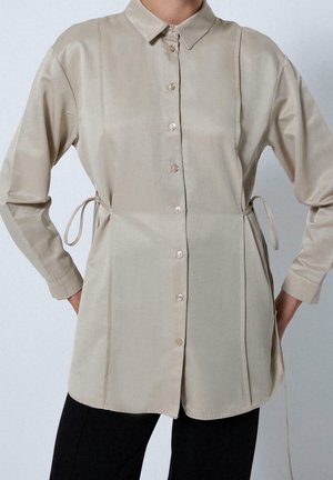 Beige langærmet skjorte med knapper, krave og sidelommer, båret over sorte bukser med hænderne hvilende på hofterne.