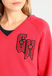 Il maglione rosso presenta un design a V con scritte ricamate in bianco e nero e accenti a forma di stella. Tessuto morbido e strutturato.