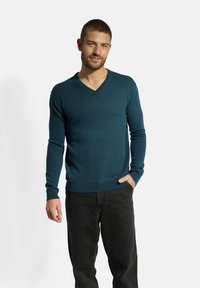 Tyrkysový svetr s výstřihem do V, vyrobený z měkké pleteniny, s dlouhými rukávy a žebrovanými manžetami, oblečený s černými kalhotami, stojící na bílém pozadí.