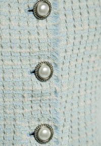 Lichtblauwe tweedstof met een gestructureerde weving, met drie parelachtige knopen met zilveren accenten aan de voorkant. Vervilte randen zichtbaar.