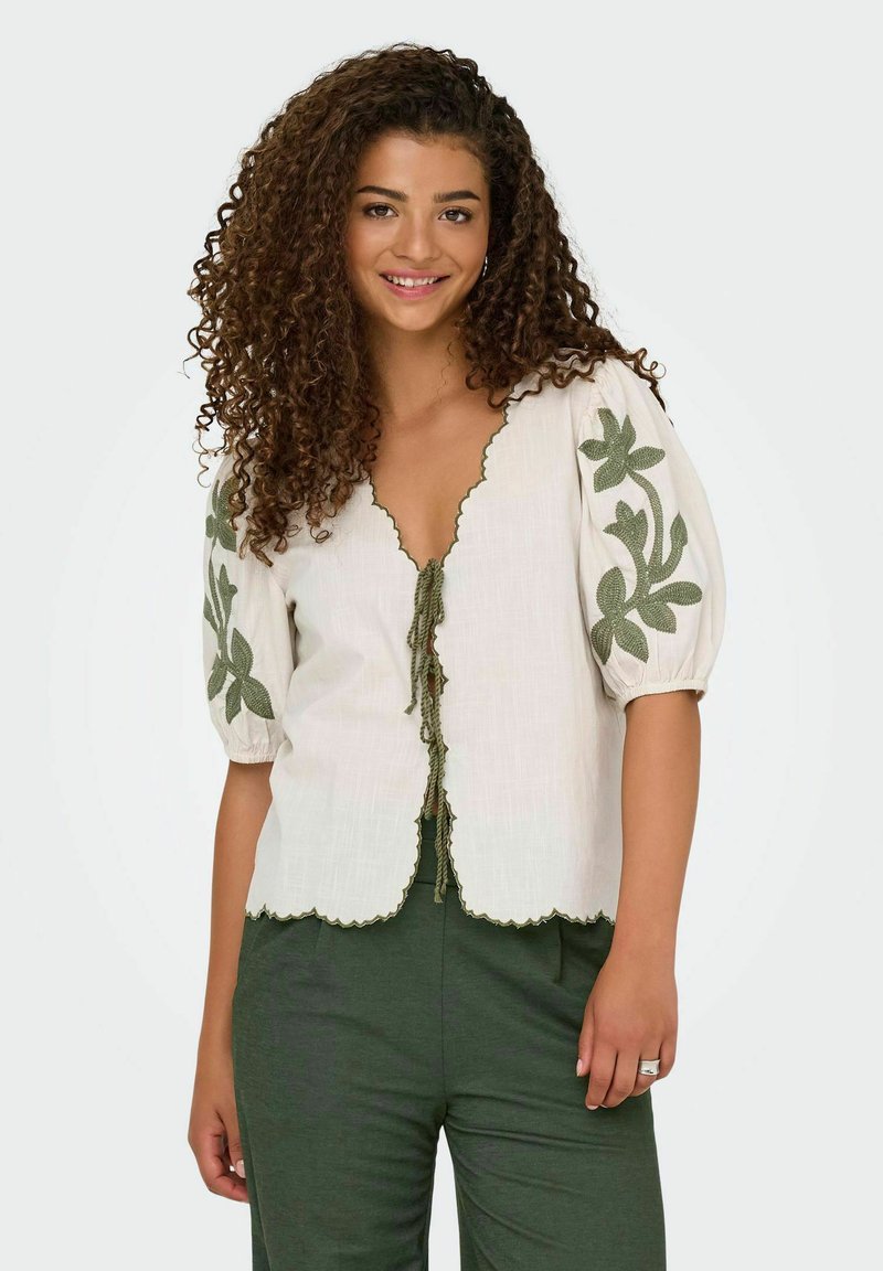 Blouse en lin blanc avec des manches bouffantes et broderie florale verte. Ourlet festonné et détail à nouer sur le devant. Associée avec un pantalon vert.