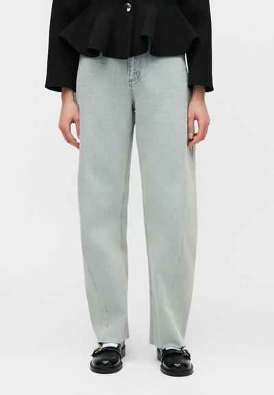 Άτομο που φοράει φαρδιά τζιν παντελόνια ανοιχτού μπλε χρώματος, μαύρο μπουφάν και μαύρα loafers με χρυσές λεπτομέρειες αγκράφας, στέκεται σε απλό φόντο.