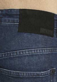 Mörkblå denimjeans med en svart tygpatch med "DRYKORN" präglat. Synlig sömnad och detaljer på bakfickan.
