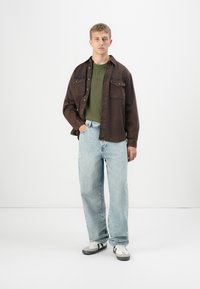 Ung mann som står, iført en mørkebrun skjorte med knapper over en olivengrønn ribbestrikket skjorte, lyseblå jeans og hvite joggesko med svarte striper.