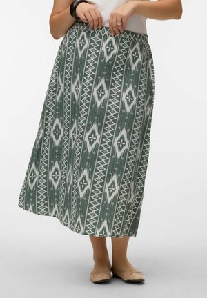 Femme portant une jupe longue à motifs verts et blancs, un haut blanc, un bracelet noir et des ballerines beige, posant devant un fond blanc.